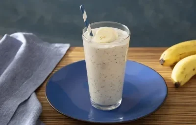 Foto da receita de Smoothie de Banana com Farinha Láctea Nestlé. Observa-se um copo com a bebida e um canudo azul e branco. Ao lado direito, um cacho de bananas