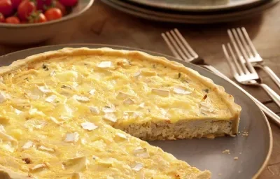 Fotografia em tons de marrom, terrosos e vermelho, com prato em tom terroso contendo uma quiche fatiada e em seu entorno três garfos, pratos empilhados com um pedaço de quiche, potinho com tomate-cereja, tudo sobre bancada de madeira.