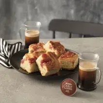 Fotografia em tons de marrom em uma bancada de madeira com um pano de prato com listra marrons, um prato redondo raso marrom com pedaços de bolo de goiabada. Ao lado, uma xícara de café Nescafé Dolce Gusto.