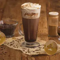 Fotografia em tons de marrom em uma bancada de madeira com um copo grande de vidro com a bebida de Alpino, chocolate quente. Ao fundo, cápsulas de alpino da Dolce Gusto, um vidrinho de canela, um copinho com raspas de chocolate.