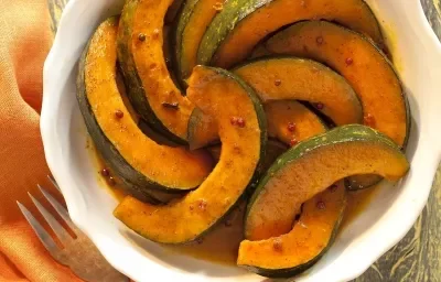 Fotografia em tons de laranja em uma bancada de madeira, ao lado um garfo apoiado em um guardanapo laranja e ao centro uma travessa redonda branca com as abóboras caramelizadas dentro.