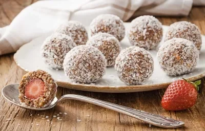 Fotografia em tons de branco, marrom e vermelho, ao centro prato branco com bombons enrolados em coco ralado, no entorno colher com bombom aberto ao meio, um morango, guardanapo branco com listras marrom, tudo sobre bancada de madeira rústica.