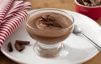 Fotografia em tons de marrom, branco e vermelho de uma bancada de madeira com um prato branco redondo, sobre ele uma taça com o mousse de chocolate zero, raspas de chocolate e uma colher. Ao lado um paninho listrado vermelho e branco.