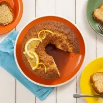 Fotografia em tons de bege, laranja, amarelo e verde de uma bancada vista de cima. Ao centro um prato redondo laranja com um bolo que contém pedaços de laranja por cima. Ao lado direito dois pratos ambos com fatias do bolo e o ao lado esquerdo também.