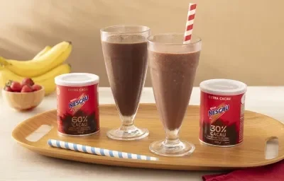 Foto em tons de vermelho da receita de shake saudável feita com Nescau Extra Cacau servida em dois copos altos de vidro sobre uma tábua de madeira com duas latas de nescau e canudos listrados. Ao fundo, bananas e morangos para decorar sobre uma mesa bege Foto em tons de vermelho da receita de shake saudável feita com Nescau Extra Cacau servida em dois copos altos de vidro sobre uma tábua de madeira com duas latas de nescau e canudos listrados. Ao fundo, bananas e morangos para decorar sobre uma mesa bege