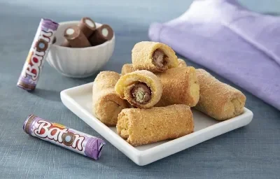Foto da receita de rolinhos de baton algodão doce servida em diversas porções sobre uma base de porcelana quadrada em cima de uma mesa acinzentada com um paninho lilás e algumas unidades de baton ao lado Foto da receita de rolinhos de baton algodão doce servida em diversas porções sobre uma base de porcelana quadrada em cima de uma mesa acinzentada com um paninho lilás e algumas unidades de baton ao lado
