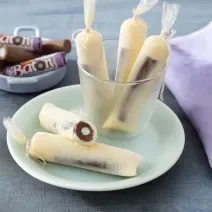 Foto da receita de geladinho algodão doce servida em 5 porções dentro de um pote transparente em cima de um prato de porcelana com um paninho lilás e algumas unidades de baton ao fundo