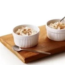Foto da receita de Arroz Doce Vegano. Observa-se uma tábua de madeira com dois potinhos brancos e pequenos de arroz doce em cima.