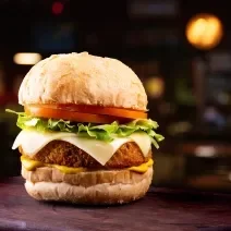 Fotografia de um hamburguer com tomate, alface, queijo, hambúrguer empanado e um molho amarelo situado ao centro de uma foto em tons escuros