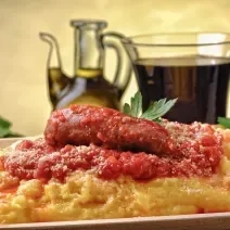 Fotografia de uma polenta com molho de tomate, cebolas e linguiça, que está dentro de um prato fundo de madeira. Por cima, queijo ralado e algumas folhas de salsinha. No fundo, muito azeite em um recipiente de vidro na frente de uma parede amarela.