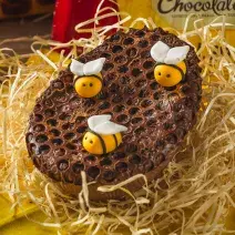 Foto aproximada de um ovo de chocolate de colher decorado com modelo de favo de mel e abelhas de mentira, sobre uma bancada com palha, tecido amarelo e uma barra de chocolate Garoto ao fundo