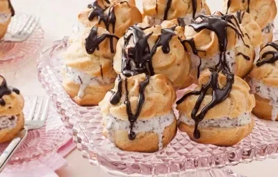 Fotografia em tons de rosa de uma bancada de rosa com um suporte de vidro com profiteroles. Ao lado dois pratos de vidro pequenos com um profiteroles e um garfo.