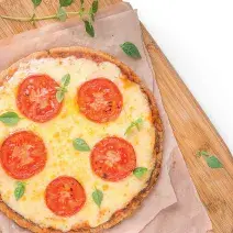 Fotografia em tons de amarelo e vermelho em uma mesa branca com uma tábua de madeira, um pano bege e uma pizza média de queijo com tomate em cima dela.