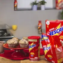 Foto vista de frente para um bancada com produtos Nescau à direita, mais ao centro um prato vermelho alta com quatro Muffins Crocantes de Nescau e à esquerda uma caneca com leite e Nescau.