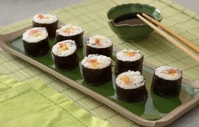 Foto em tons de verde da receita de hossomaki básico servida em 9 porções dispostas sobre uma régua de porcelana verde escura com borda marrom. Ao lado há dois hashis sobre um recipiente verde escuro com shoyo