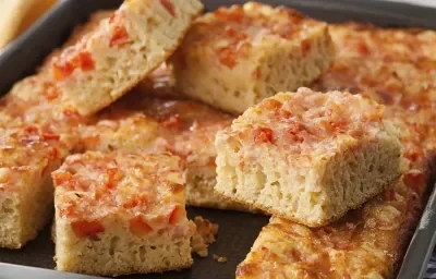 Fotografia em tons de marrom e vermelho em uma bancada de madeira com um recipiente retangular com vários pedacinhos de torta de bauru.