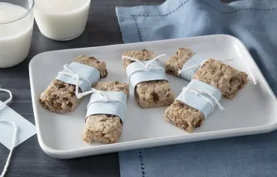 Foto em tons de azul claro da receita de barrinha de iogurte nesfit e aveia servida em 5 porções sobre uma régua de porcelana branca sobre uma mesa cinza com um pano azul ao fundo além de dois copos com iogurte Foto em tons de azul claro da receita de barrinha de iogurte nesfit e aveia servida em 5 porções sobre uma régua de porcelana branca sobre uma mesa cinza com um pano azul ao fundo além de dois copos com iogurte