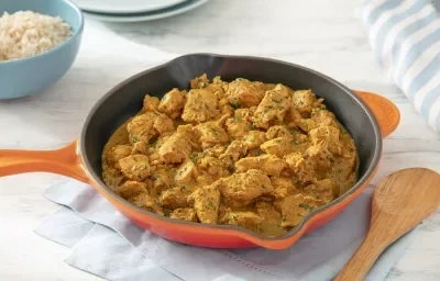 Foto da receita de frango ao curry sem lactose servida em uma frigideira grande laranja sobre uma mesa clara com um pano azul embaixo e uma colher de madeira ao lado