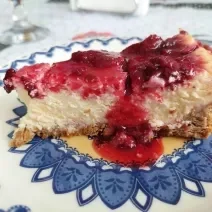 Foto em tons de vermelho da receita de cheesecake de frutas vermelhas ao estilo nova york servida em uma fatia sobre um prato decorado de porcelana azul e branco