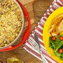 Fotografia em tons de amarelo e vermelho em uma bancada de madeira com um pano vermelho listrado com um prato amarelo com salada verde, arroz, frango e farofa de feijão. Ao lado, uma frigideira vermelha com farofa dentro.