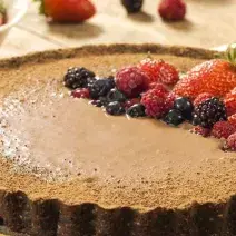 Fotografia em tons de marrom em uma mesa com uma toalha bege, um prato raso redondo grande com a torta de biscoito Nesfit com cacau e frutas vermelhas para decorar a torta, em cima do prato.