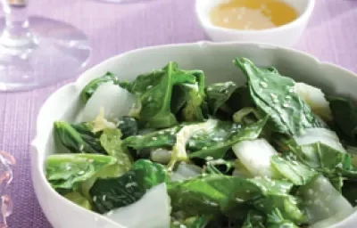 Fotografia em tons de branco, verde e lilás, ao centro uma saladeira com folhas verdes e gergelim, ao lado potinho de azeite, copos e uma jarra, sobre uma toalha lilás,