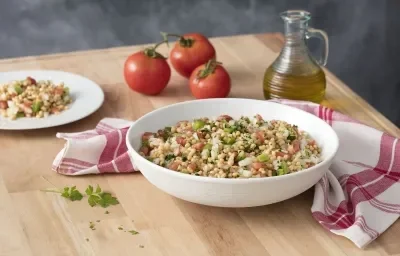 Fotografia em tons de branco e vermelho, ao centro recipiente branco com salada de feijão e tomate, ladeada por prato com salada, três tomates, galheteiro com azeite e guardanapo branco com listras vermelhas, tudo sobre bancada de madeira e fundo cinza.