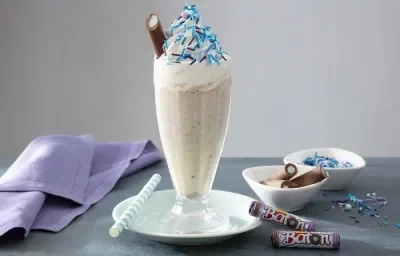 Foto da receita de milkshake algodão doce servida em um copo largo e alto de vidro sobre uma mesa acinzentada com um paninho lilás ao lado, além de algumas unidades de baton