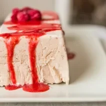 Fotografia de uma terrine de iogurte, em formato de uma forma retangular. Por cima, uma calda de frutas vermelhas, sobre um prato branco quadrangular.