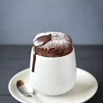 Fotografia de um suflê de café com NESCAFÉ e açúcar de confeiteiro polvilhado por cima. A sobremesa está em um recipiente de vidro branco alto em cima de um prato pequeno branco de sobremesa, e uma colher de café apoiada.