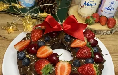 Foto da receita de guirlanda de brownie decorada com frutas vermelhas e decoração de natal servida em um prato grande branco sobre uma mesa de madeira com produtos de leite moça ao fundo