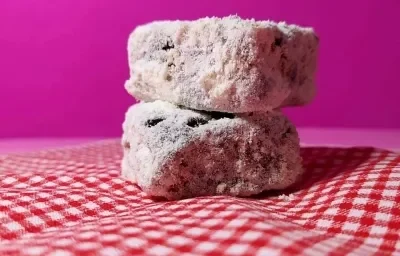 Foto da receita de Palha Italiana. Observa-se duas palhas italianas cortadas em quadrados dispostas em uma toalha quadriculada branca e vermelha e em um fundo rosa.