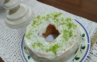 Foto em tons de branco da receita de bolo de nescafé com cobertura de limão servida em uma porção grande sobre um prato decorado em cima de uma mesa de madeira com um pano bordado