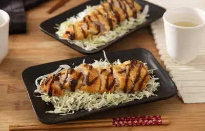 Foto da receita de Tonkatsu (Bife de Porco Japonês). Observa-se dois pratos retangulares pretos com repolho fatiado e o tonkatsu por cima, servido com um pouco de molho escuto. Observa-se um hashi na foto, na frente do prato.