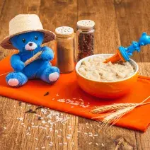 Fotografia em tons de laranja e azul de uma bancada de madeira com um paninho laranja e um pote laranja com a papinha. Na frente, grãos de arroz e cravos decorando e uma pelúcia azul com chapéu de palha.