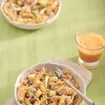 Fotografia em tons de verde em uma bancada de madeira verde com um pano bege, um prato fundo bege com a salada colorida com macarrão, ao lado um copo de vidro com um suco de laranja.