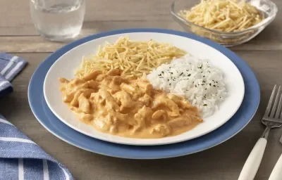 Fotografia em tons de azul em uma bancada de madeira com um sousplat azul, um prato branco raso em cima com o strogonoff, arroz branco e batata palha. Ao lado, um pano azul com listras brancas, talheres, copo com água e um potinho com batata palha. Fotografia em tons de azul em uma bancada de madeira com um sousplat azul, um prato branco raso em cima com o strogonoff, arroz branco e batata palha. Ao lado, um pano azul com listras brancas, talheres, copo com água e um potinho com batata palha.