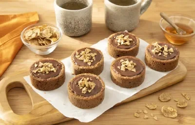 Foto da Receita de Torta de Banana e Cacau com Nesfit Cereal. Observa-se uma tábua de madeira com 6 tortinhas sobre ela, recheadas de chocolate e polvilhas de cereal nesfit por cima. Cereais e um potinho com mel decoram a foto. Foto da Receita de Torta de Banana e Cacau com Nesfit Cereal. Observa-se uma tábua de madeira com 6 tortinhas sobre ela, recheadas de chocolate e polvilhas de cereal nesfit por cima. Cereais e um potinho com mel decoram a foto.