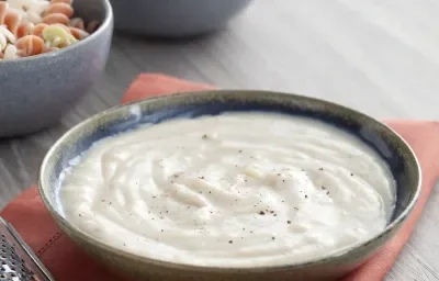 Fotografia em tons de cinza e vermelho de uma bancada de madeira cinza com um paninho vermelho, sobre ele um recipiente redondo cinza com molho branco, uma noz moscada e um ralador. Ao fundo dois recipientes cinzas com macarrão colorido e molho branco.