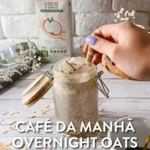 Foto da receita de overnight oats servida em um recipiente hermético alto com uma mão com uma colher, isso sobre uma mesa de madeira cinza. Ao fundo, há uma embalagem de bebida vegetal de coco e arroz antures heart