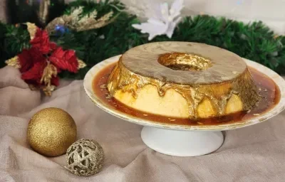 Fotografia em tons de dourado em uma bancada com um pano dourado e enfeites de natal. Ao centro uma bandeja redonda com um pudim de leite Moça e uma calda de caramelo com corante dourado comestível em cima.