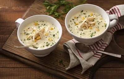 Fotografia em tons de marrom em uma mesa de madeira escura com uma tábua de madeira ao centro e dois pratos fundos com a sopa cremosa de milho com frango dentro delas. Ao lado, um guardanapo bege com listras vermelhas.