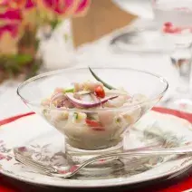 Fotografia em tons de branco e vermelho de uma mesa branca com um prato vermelho, sobre ele um prato branco florido, uma colher prata, e uma taça com pedaços de peixe, cebola roxa, pimenta e cebolinha. Ao fundo um arranjo de flores.