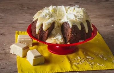 Fotografia em tons de amarelo, em uma bancada de madeira de cor marrom. Ao centro, um pano amarelo contendo o bolo vulcão. Ao lado, há 3 pedaços de chocolate branco e algumas raspas de chocolate espalhadas.