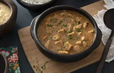 Foto da receita de Sarapatel. Observa-se uma panela de barro com a receita sobre uma tábua de madeira em uma superfície preta. Em cima, cumbucas de arroz e farofa.