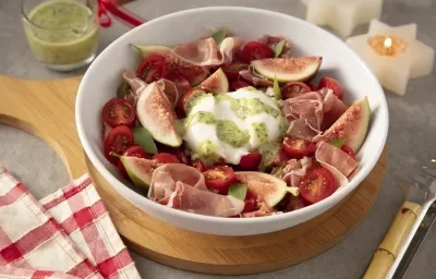 Foto da receita de Burrata ao Pesto com Presunto Cru e Figos. Observa-se um recipiente refratário branco de porcelana redondo com a burrata dentro ao centro, e em volt os presuntos, figos e tomates. Observa-se molho pesto regado por cima. Foto da receita de Burrata ao Pesto com Presunto Cru e Figos. Observa-se um recipiente refratário branco de porcelana redondo com a burrata dentro ao centro, e em volt os presuntos, figos e tomates. Observa-se molho pesto regado por cima.