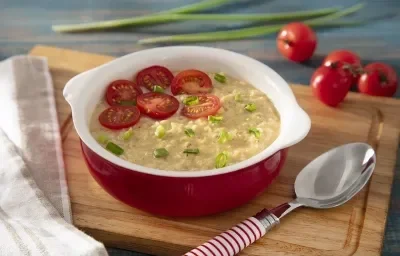 Foto em tons de vermelho da receita de aveia salgada servida em uma panelinha de cerâmica vermelha sobre uma tábua de madeira com uma colher prateada ao lado. Ao fundo temos alguns tomates-cereja