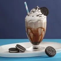 Foto da receita de Milkshake de negresco. Observa-se um copo de milkshake de vidro transparente decorado com calda e com a bebida dentro e topping de chantilly e farofa de biscoito, com um canudo azul e branco espetado