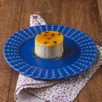 imagem da receita de pudim de iogurte da rafaela masaia, servido em um prato azul com uma calda de frutas amarelas. O prato está sobre um pano florido em tom rosa em uma bancada de madeira