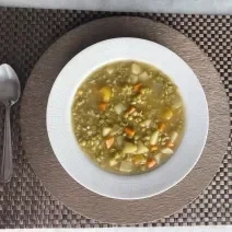 Imagem da receita de Sopa de Ervilha com Legumes, em um prato branco, sobre uma mesa e ao lado uma colher